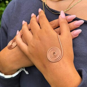 May include: Copper spiral jewelry set. The set includes a bracelet and a ring, both featuring a coiled spiral design. The jewelry is a warm copper color and is worn on a person's hands. The background is a blurred green and blue.