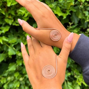 May include: Two hands each adorned with a copper-colored spiral bracelet. The bracelets are made of thin wire, with a spiral design on the palm and a connecting band. The background is a lush green with leafy foliage.