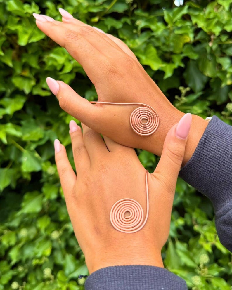 May include: Two hands each adorned with a copper-colored spiral bracelet. The bracelets are made of thin wire, with a spiral design on the palm and a connecting band. The background is a lush green with leafy foliage.