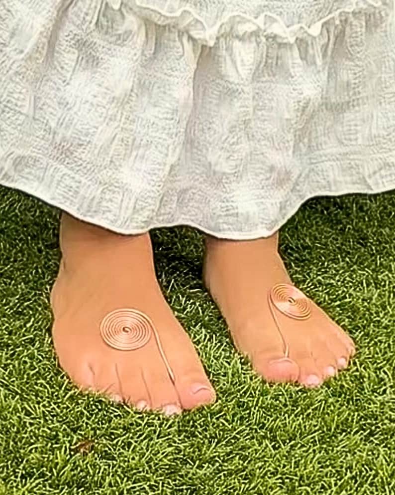 May include: A pair of feet with copper-colored toe rings, each featuring a spiral design. The feet are on a bed of green grass, with a white dress visible in the background.