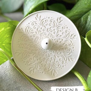 Puede incluir: Un objeto decorativo blanco y circular con un intrincado diseño floral simétrico. Presenta un pequeño pomo blanco en el centro y el texto "DESIGN B" en la esquina inferior derecha. El objeto se encuentra sobre un fondo de follaje verde.
