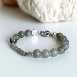 May include: A beaded bracelet featuring gray and silver-toned beads. The bracelet has a mix of small and large round beads, with a silver clasp. The beads have a speckled appearance. The bracelet is on a white surface.