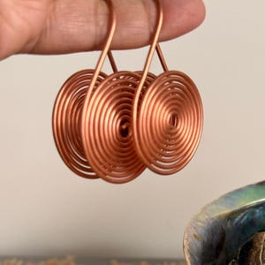 May include: Three copper-colored spiral earrings. Each earring features a circular design with concentric coils. The earrings are held up against a neutral background, showcasing their metallic sheen and unique shape.