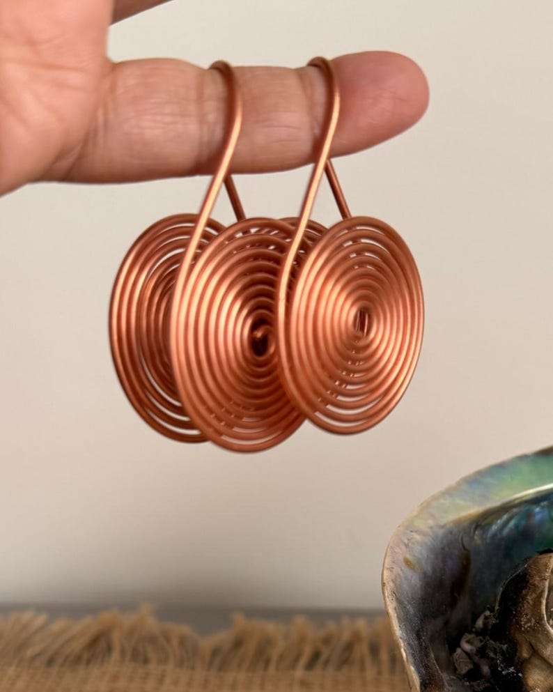May include: Three copper-colored spiral earrings. Each earring features a circular design with concentric coils. The earrings are held up against a neutral background, showcasing their metallic sheen and unique shape.