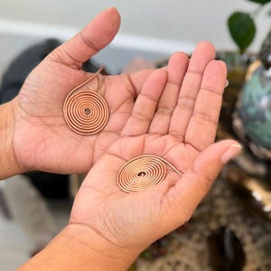 May include: Two copper spiral pendants, each held in an open palm. The pendants are made of thin, coiled copper wire, forming a circular shape. The background is blurred, with hints of green foliage.