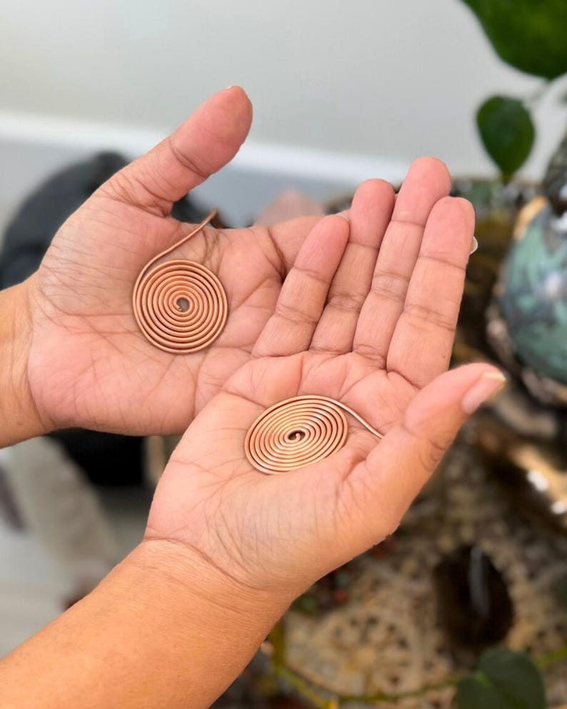May include: Two copper spiral pendants, each held in an open palm. The pendants are made of thin, coiled copper wire, forming a circular shape. The background is blurred, with hints of green foliage.
