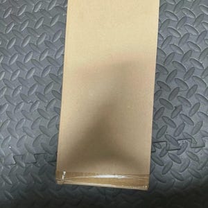 May include: A rectangular cardboard box with a light brown, tan finish. The box is oriented vertically and features a single strip of clear adhesive tape securing the bottom edge, providing a clean, plain surface for shipping or storage purposes.