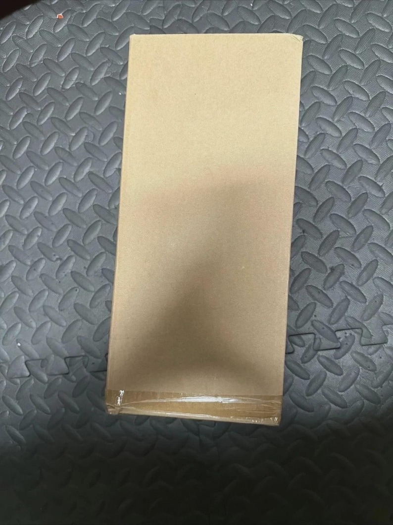 May include: A rectangular cardboard box with a light brown, tan finish. The box is oriented vertically and features a single strip of clear adhesive tape securing the bottom edge, providing a clean, plain surface for shipping or storage purposes.