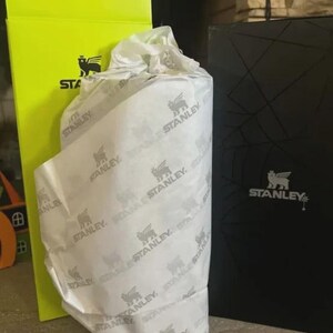 May include: A cylindrical object wrapped in white tissue paper printed with the black Stanley logo and winged bear icon, positioned between a tall, bright neon yellow box and a black box, both featuring the Stanley brand name and logo.