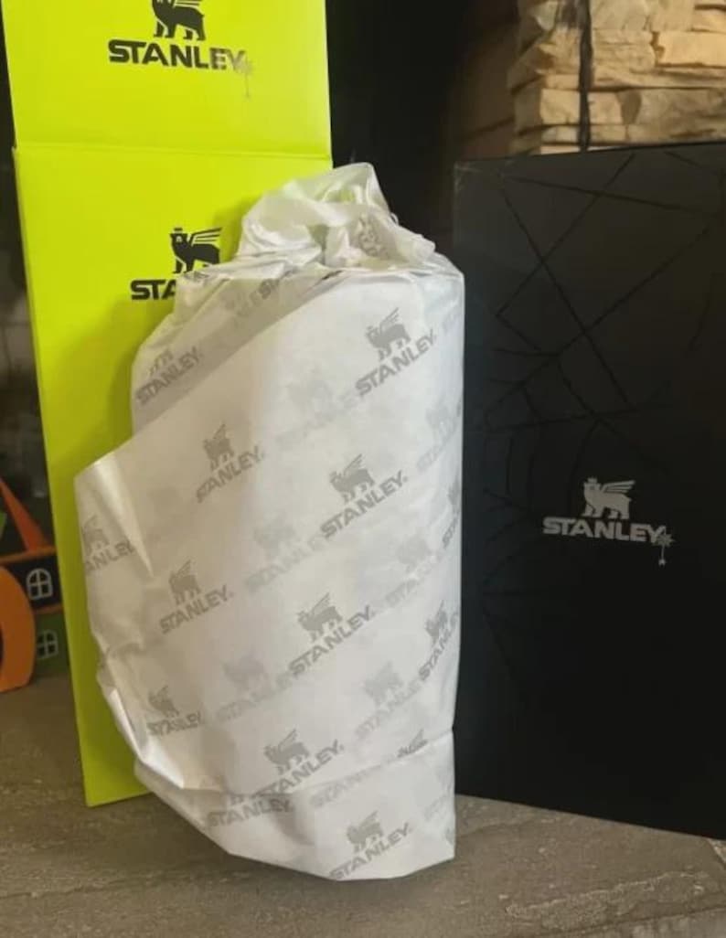 May include: A cylindrical object wrapped in white tissue paper printed with the black Stanley logo and winged bear icon, positioned between a tall, bright neon yellow box and a black box, both featuring the Stanley brand name and logo.