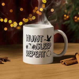 May include: White ceramic mug featuring the text "HUNT SLEEP REPEAT" in black, with a bird and dog silhouette. The mug is on a wooden surface, with cinnamon sticks visible.