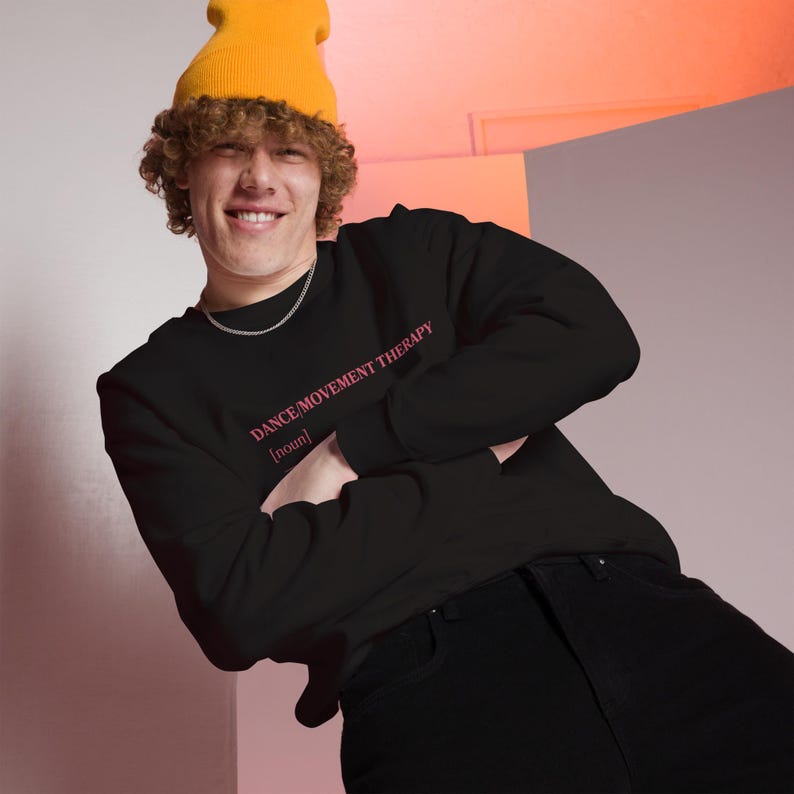 May include: Black sweatshirt with the text "DANCE/MOVEMENT THERAPY [noun]" in pink. The person is wearing a yellow beanie and a silver chain necklace. The person is wearing black jeans.