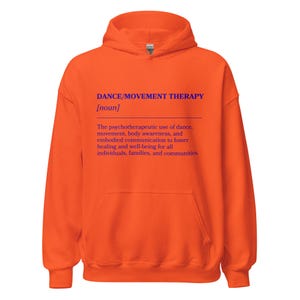 May include: An orange hoodie with a front pocket and a hood. The front of the hoodie features the text "DANCE/MOVEMENT THERAPY [noun]" in blue, along with a definition of the term. The hoodie is made of a soft material.