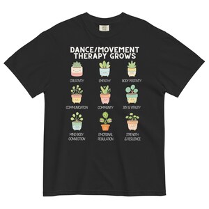 May include: Black t-shirt with the text "DANCE/MOVEMENT THERAPY GROWS" and illustrations of potted plants. Each plant represents a concept like creativity, empathy, and joy & vitality. A comfortable, casual tee.