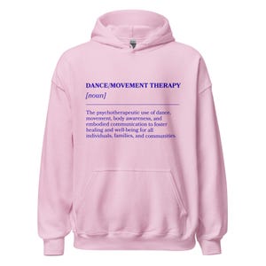 May include: A light pink hoodie with a drawstring hood and a kangaroo pocket. The front of the hoodie features the text "DANCE/MOVEMENT THERAPY [noun]" in purple, along with a definition of the term.