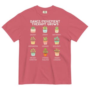 May include: A red t-shirt with the text "DANCE/MOVEMENT THERAPY GROWS" and illustrations of potted plants. Below the text are the words "CREATIVITY", "EMPATHY", "BODY POSITIVITY", "COMMUNICATION", "COMMUNITY", "JOY & VITALITY", "MIND BODY CONNECTION", "EMOTIONAL REGULATION", and "STRENGTH & RESILIENCE".