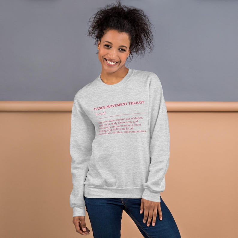 May include: A light gray sweatshirt with the text "DANCE/MOVEMENT THERAPY" in red. The definition of dance/movement therapy is printed below the title. The person in the image is wearing blue jeans.