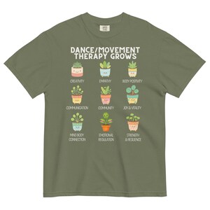 May include: Olive green t-shirt with the text "DANCE/MOVEMENT THERAPY GROWS" above illustrations of potted plants. Each plant represents a positive concept like creativity, empathy, and joy & vitality.