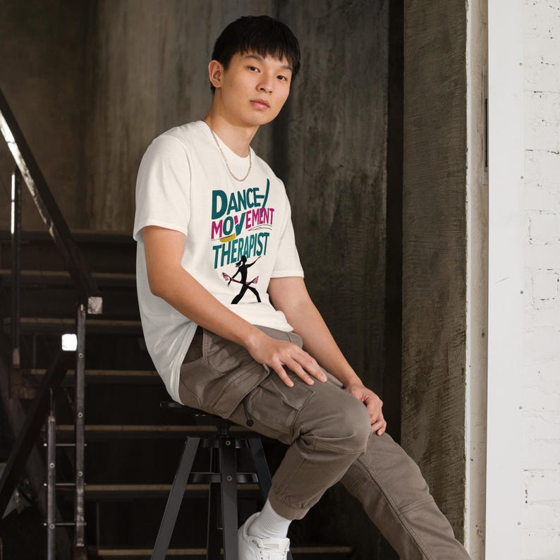 May include: Off-white t-shirt with the text "DANCE MOVEMENT THERAPIST" in various colors and a silhouette of a dancer. The shirt is short-sleeved.