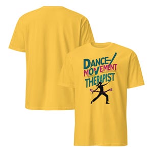 May include: A yellow t-shirt with the text "DANCE MOVEMENT THERAPIST" in teal, pink, and yellow. A black silhouette of a person dancing with two purple batons is below the text. Another yellow t-shirt is visible in the background.