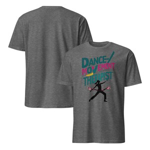 May include: A heather gray t-shirt featuring the text "DANCE/MOVEMENT THERAPIST" in teal, pink, and yellow. A black silhouette of a person dancing with pink props is below the text. Another gray t-shirt is visible in the background.