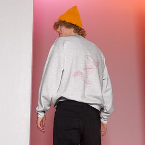 May include: A light gray sweatshirt with a pink graphic design on the back. The design appears to be made up of several words. The person in the image is wearing a yellow beanie and black pants.