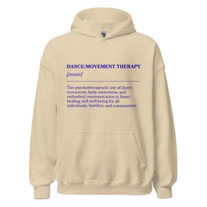 May include: A beige hoodie with a drawstring hood and a front pocket. The front of the hoodie features the text "DANCE/MOVEMENT THERAPY [noun]" in blue, along with a definition of the term.