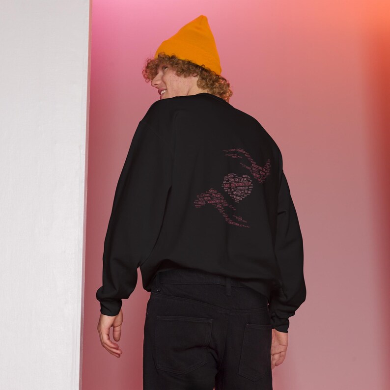 May include: Black sweatshirt with a heart and hand design in pink. The sweatshirt is worn with black jeans and a yellow beanie. The design on the back of the sweatshirt is made up of words.