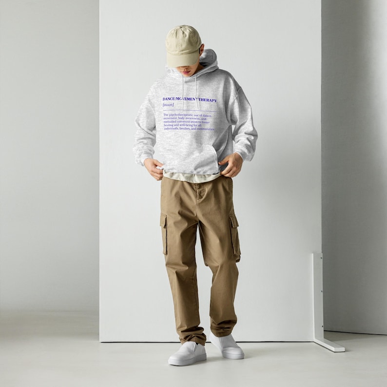 May include: A light gray hoodie with the text "DANCE MOVEMENT THERAPY" in blue, along with a definition. The person is wearing khaki cargo pants, a tan baseball cap, and white sneakers.