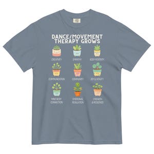 May include: A blue t-shirt with the text "DANCE/MOVEMENT THERAPY GROWS" and illustrations of potted plants representing concepts like creativity, empathy, and joy. The shirt is a comfortable, casual wear item.