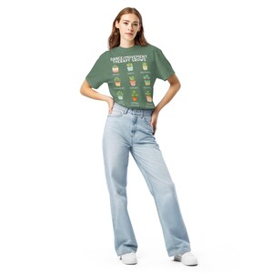 May include: A green t-shirt with the text "DANCE/MOVEMENT THERAPY GROWS" and illustrations of potted plants. The model is wearing light blue jeans and white sneakers.