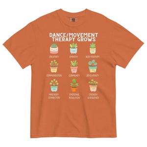 May include: Orange t-shirt with the text "DANCE/MOVEMENT THERAPY GROWS" and illustrations of potted plants. Each plant represents a concept like creativity, empathy, and joy. The shirt is a comfortable fit.