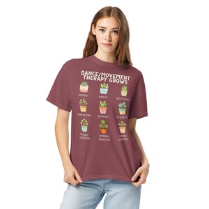 May include: A maroon t-shirt with the text "DANCE/MOVEMENT THERAPY GROWS" and illustrations of potted plants. Each plant represents a positive aspect of therapy, such as creativity, empathy, and body positivity. The shirt is worn by a person.