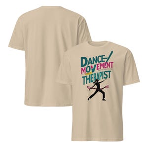 May include: Beige t-shirt featuring the text "Dance Movement Therapist" in teal, pink, and yellow. A black silhouette of a dancer with pink ribbons is below the text. Another beige t-shirt is visible in the background.