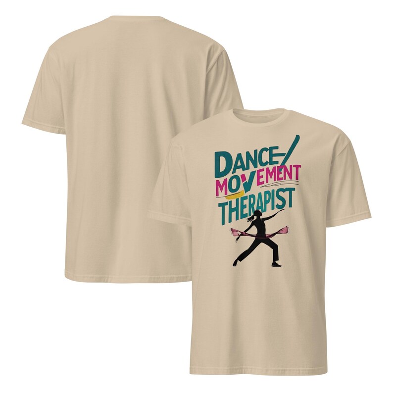 May include: Beige t-shirt featuring the text "Dance Movement Therapist" in teal, pink, and yellow. A black silhouette of a dancer with pink ribbons is below the text. Another beige t-shirt is visible in the background.