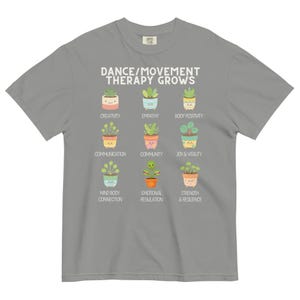 May include: A gray t-shirt with the text "DANCE/MOVEMENT THERAPY GROWS" in white. Below the text are nine colorful illustrations of potted plants, each labeled with a positive concept like "Creativity" and "Empathy".