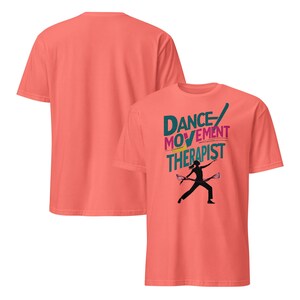 May include: Coral-colored t-shirts. One shirt displays the text "DANCE MOVEMENT THERAPIST" in teal, pink, and yellow, with a black silhouette of a person holding two objects. The second shirt is a solid coral color.