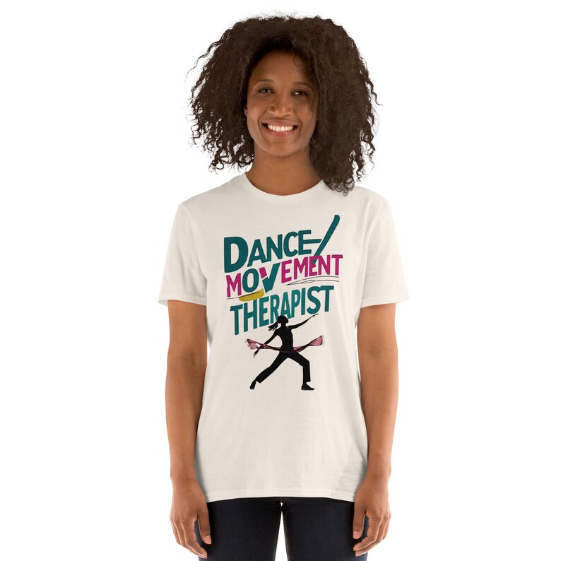May include: A cream-colored t-shirt with the words "DANCE MOVEMENT THERAPIST" in teal, pink, and yellow. A silhouette of a person dancing with two pink batons is below the text. The shirt is a short-sleeved crew neck.