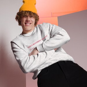 May include: A light gray sweatshirt with the text "DANCE/MOVEMENT THERAPY [noun]" in red. The sweatshirt is worn with a yellow beanie and black pants. The person is smiling with arms crossed.