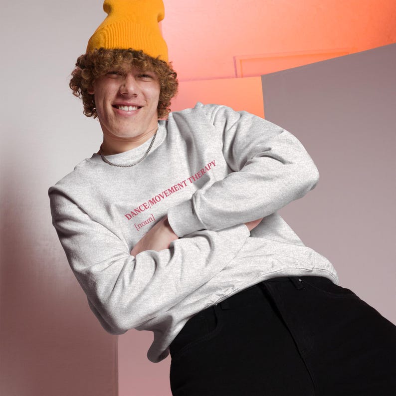 May include: A light gray sweatshirt with the text "DANCE/MOVEMENT THERAPY [noun]" in red. The sweatshirt is worn with a yellow beanie and black pants. The person is smiling with arms crossed.