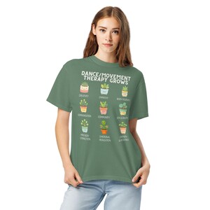 May include: A green t-shirt with the text "DANCE/MOVEMENT THERAPY GROWS" and illustrations of potted plants. Each plant represents a positive concept like creativity, empathy, and body positivity. The shirt is made of a soft material.