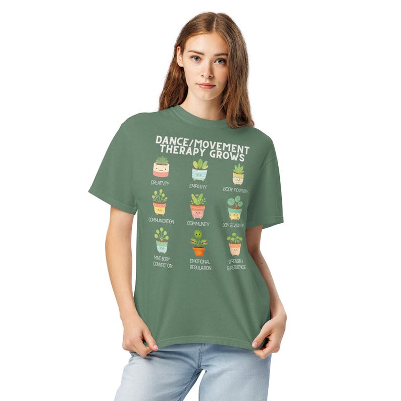 May include: A green t-shirt with the text "DANCE/MOVEMENT THERAPY GROWS" and illustrations of potted plants. Each plant represents a positive concept like creativity, empathy, and body positivity. The shirt is made of a soft material.