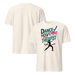May include: Off-white t-shirt featuring the text "DANCE MOVEMENT THERAPIST" in teal, pink, and yellow. A black silhouette of a dancer is in the center. The back of the shirt is plain off-white.