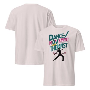 May include: A white t-shirt featuring the text "DANCE MOVEMENT THERAPIST" in teal, pink, and yellow. A black silhouette of a dancer is below the text, holding pink ribbons. The back of the shirt is plain white.