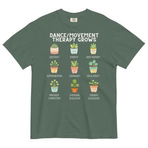 May include: A green t-shirt with the text "DANCE/MOVEMENT THERAPY GROWS" and illustrations of potted plants. Each plant represents a concept like creativity, empathy, and joy & vitality. The shirt is made of a soft material.
