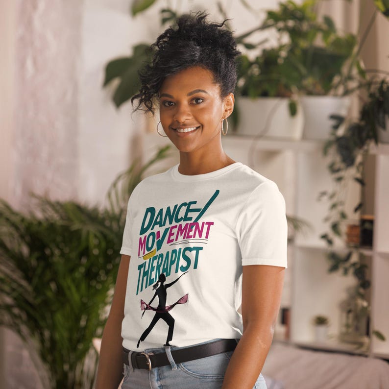 May include: White t-shirt with the text "DANCE MOVEMENT THERAPIST" in teal, pink, and yellow. A silhouette of a dancer with ribbons is below the text. The shirt is worn by a person.