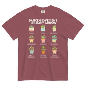 May include: A maroon t-shirt featuring the text "DANCE/MOVEMENT THERAPY GROWS" above illustrations of potted plants. Each plant represents a concept like creativity, empathy, and joy & vitality. The shirt is made of a soft, comfortable material.