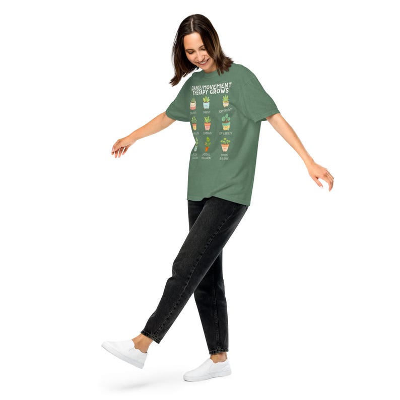 May include: A green t-shirt with the text "DANCE/MOVEMENT THERAPY GROWS" and illustrations of various potted plants. The person is wearing black jeans and white shoes.
