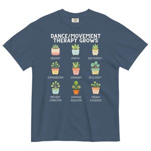 May include: A navy blue t-shirt with the text "DANCE/MOVEMENT THERAPY GROWS" and illustrations of potted plants. Each plant represents a positive concept like creativity, empathy, and joy & vitality. The shirt is made of a soft, comfortable material.