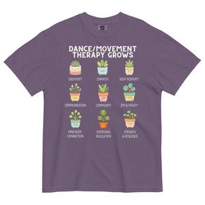 May include: A purple t-shirt with the text "DANCE/MOVEMENT THERAPY GROWS" and illustrations of potted plants representing concepts like creativity, empathy, and joy. The shirt is a comfortable, casual wear item.